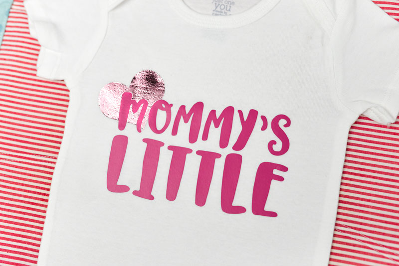 Image of a white onesie in the beginning steps of applying iron on and iron on foil.  Heart is in pink foil and the text \'Mommy\'s Little\' is in pink iron on vinyl.
