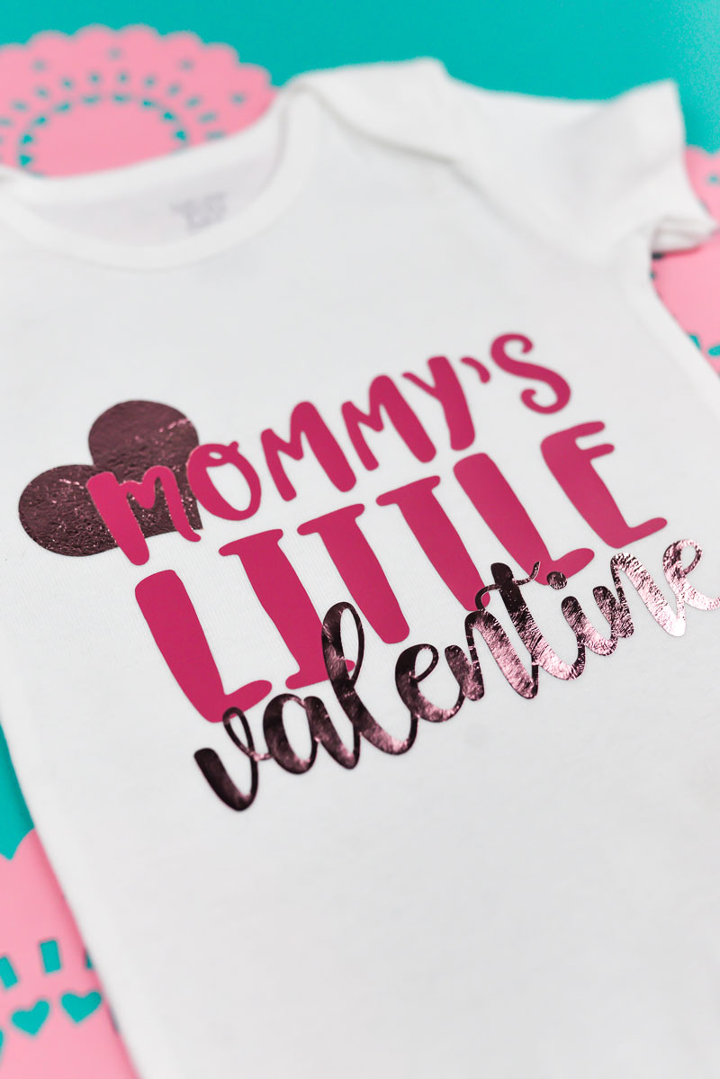 Image of a white onesie in the final steps of applying iron on and iron on foil.  Heart and the text \'valentine\' is in pink foil and the text \'Mommy\'s Little\' is in pink iron on vinyl